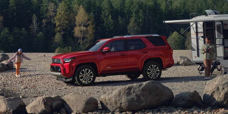Red SUV parked on a rocky shoreline next to an RV, with a woman taking photos and another running toward the vehicle