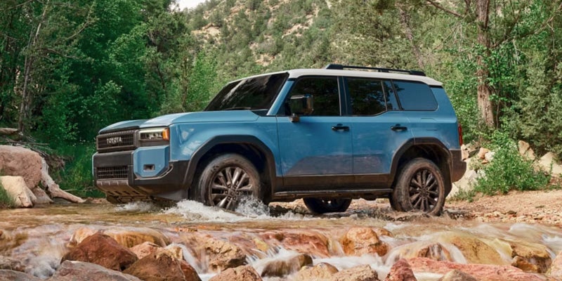 A blue Toyota off-road SUV with a white roof traversing a shallow, rocky river surrounded by lush green trees