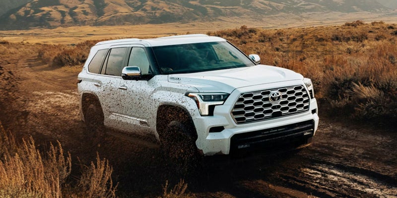 A muddy white Toyota Sequoia SUV drives on a dirt track through dry fields with mountains in the background