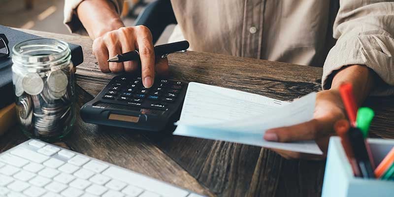 A person uses a calculator with one hand and holds a paper bill in the other at a wooden desk