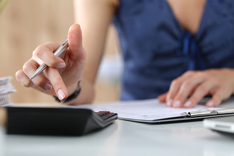 A person's hand, holding a pen, points to a calculator while paperwork and another hand are in the background.
