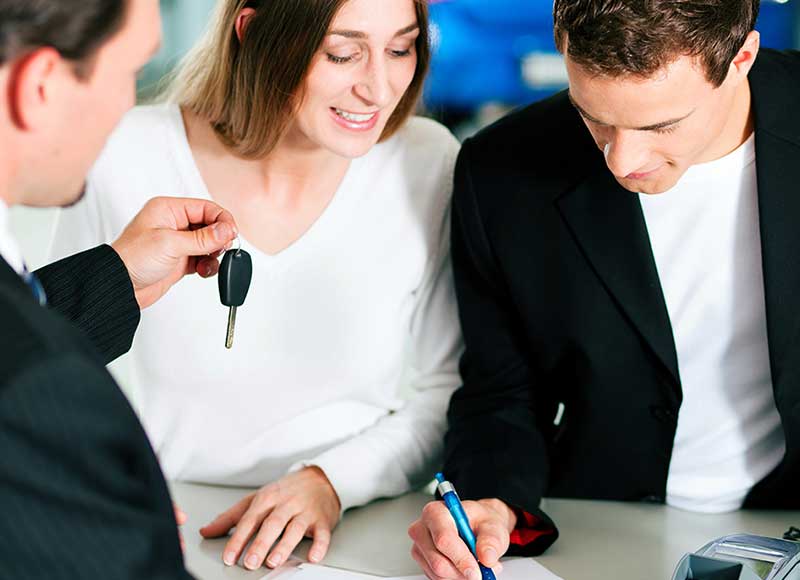 Two people sit at a desk reviewing and signing paperwork while a third person extends a car key