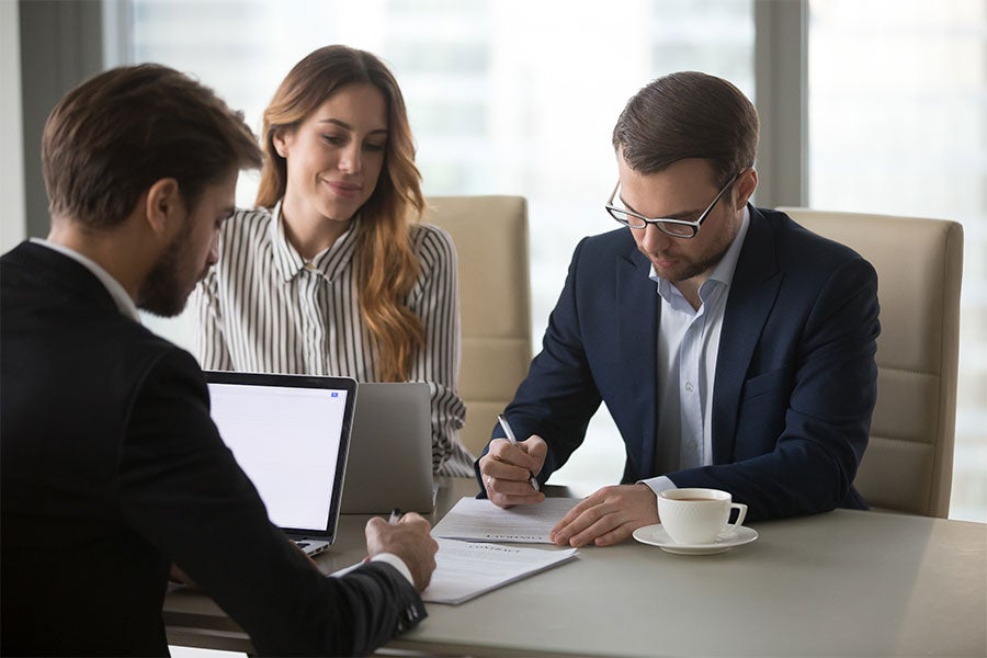 Three business professionals signing documents and using a laptop at a meeting table.