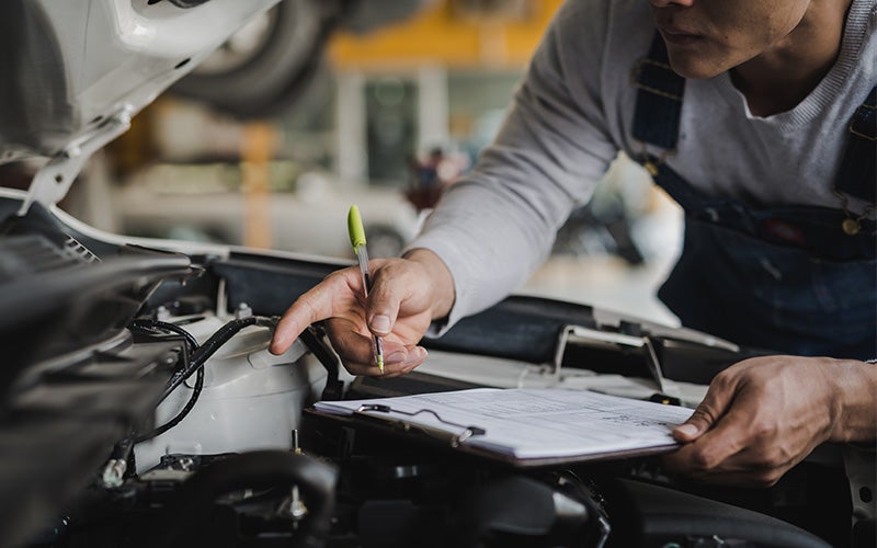 A mechanic in overalls inspects a car engine with a pen and clipboard, pointing to a component.