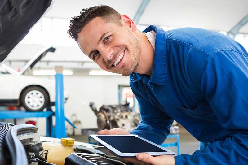 A smiling male mechanic in blue coveralls holds a tablet while leaning over an open car engine in a garage.