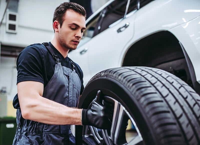Automotive technician holding and inspecting a car tire next to a white vehicle in a workshop