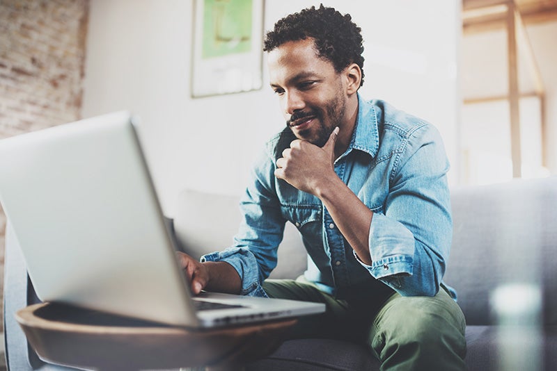 Smiling man with curly hair and beard, wearing a denim shirt, using a laptop while sitting on a couch.